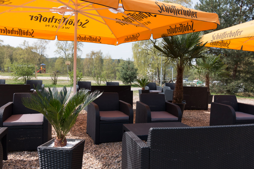 Sitzbereich im Freien mit schwarzen Korbstühlen und Tischen unter großen gelben Schöfferhofer-Sonnenschirmen, umgeben von kleinen Palmen und Kiesboden.