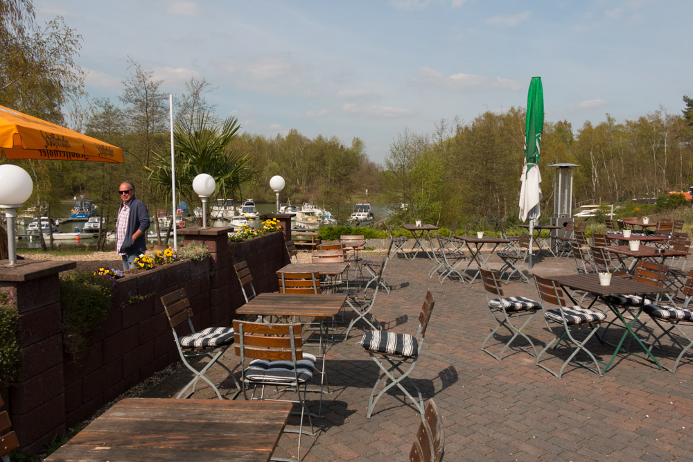 Terrasse im Freien mit leeren Tischen und Stühlen, Topfblumen und einem Mann, der am Rand entlang geht. Im Hintergrund sind Boote und Bäume unter einem teilweise bewölkten Himmel zu sehen.