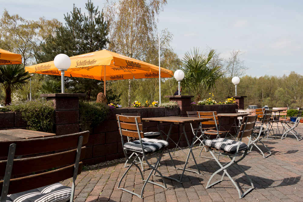 Caféterrasse im Freien mit leeren Holzstühlen und -tischen, Topfpflanzen, gestreiften Kissen und gelben Sonnenschirmen an einem sonnigen Tag. Im Hintergrund sind Bäume und Grünflächen zu sehen.