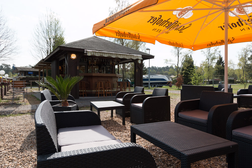 Sitzbereich im Freien mit Korbstühlen und Tischen unter einem großen orangefarbenen Sonnenschirm in der Nähe einer hölzernen Bar, umgeben von Bäumen und Grünflächen.