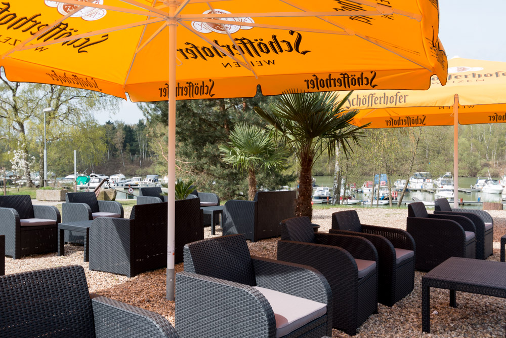 Freisitz mit Korbstühlen und kleinen Tischen unter großen orangefarbenen Sonnenschirmen, in der Nähe eines Yachthafens mit Booten und umgeben von Bäumen.