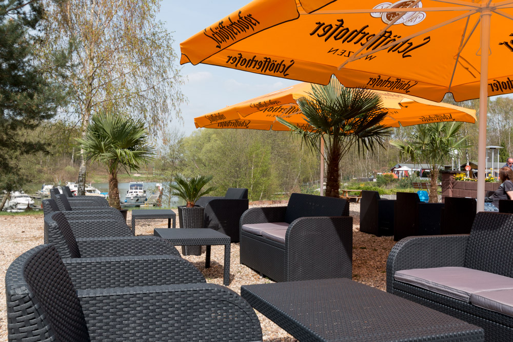 Sitzbereich im Freien mit schwarzen Korbstühlen und Tischen unter großen gelben Sonnenschirmen, umgeben von Palmen und Grünpflanzen, mit Booten im Hintergrund.
