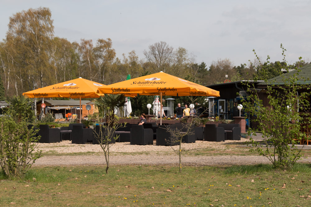 Sitzplatz im Freien mit schwarzen Möbeln unter zwei großen gelben Sonnenschirmen, umgeben von Bäumen und Sträuchern, an einem teilweise bewölkten Tag.