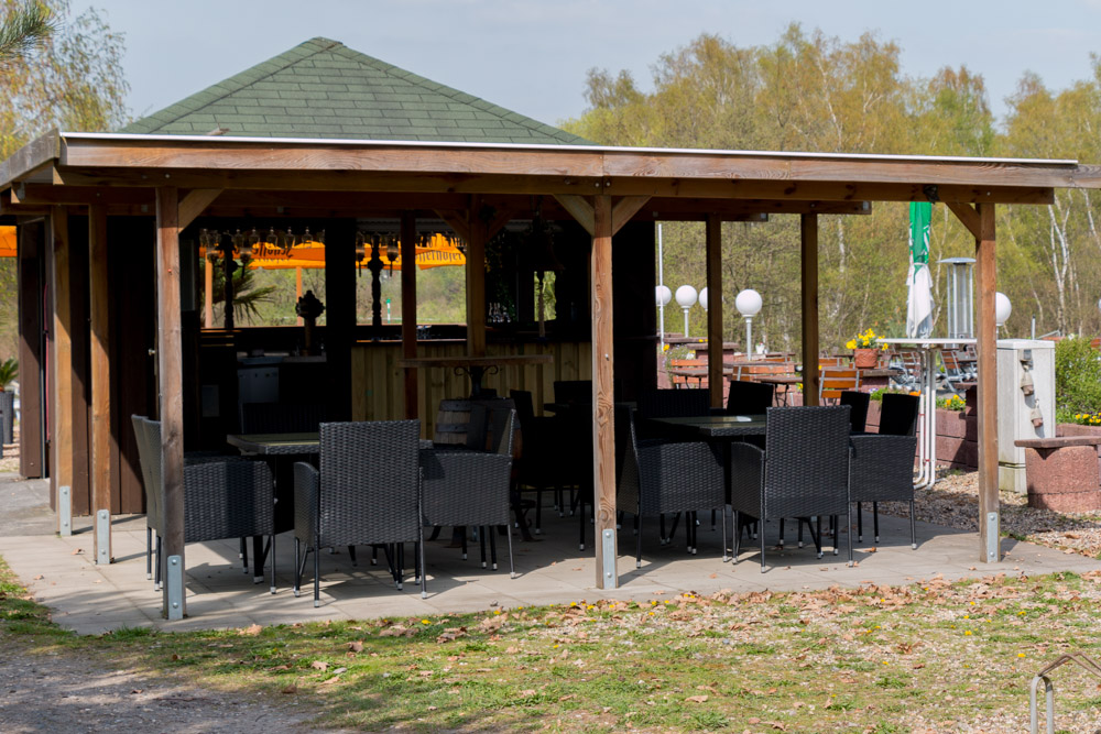 In einem hölzernen Pavillon mit grünem Dach stehen mehrere schwarze Korbtische und -stühle auf einer Steinterrasse, umgeben von Bäumen und Grünpflanzen.