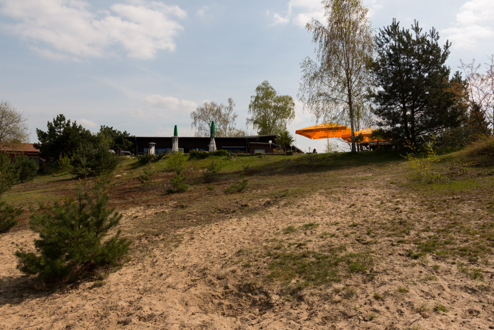 Ein sandiger Abhang mit verstreuten kleinen Büschen führt hinauf zu einem Gebäude mit Sonnenschirmen und Bäumen im Hintergrund unter einem teilweise bewölkten Himmel.